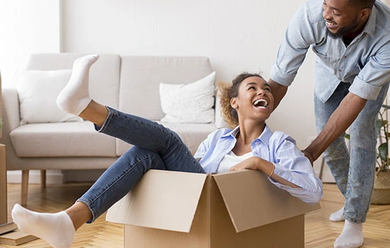 Father and daughter playing together with cardboard boxes in living room, child sitting in box with legs up while father helps lift it