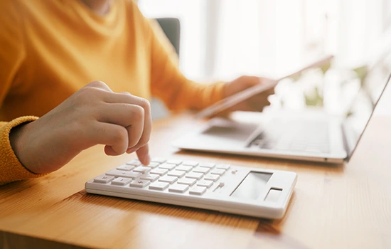 Person in orange shirt typing on wireless keyboard at wooden desk with laptop and notebook in background