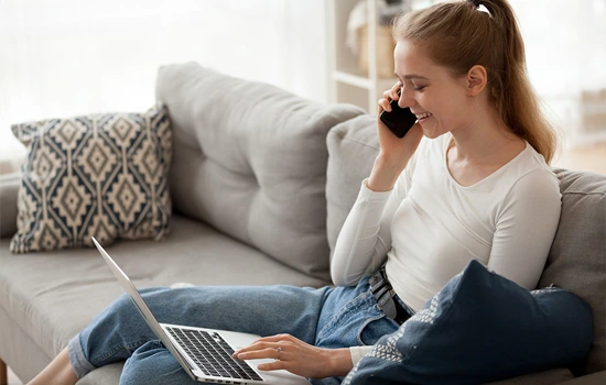 Woman sitting on gray couch with laptop, talking on phone while working from home
